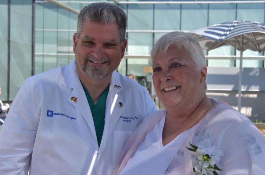 Man in white Duke labcoat leans into woman for a photo with glass building in the background