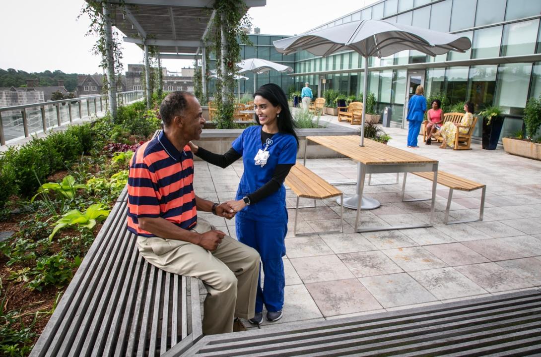 At rooftop garden, man sits on a bench while woman dressed in blue with Duke ID puts her arm on his shoulder