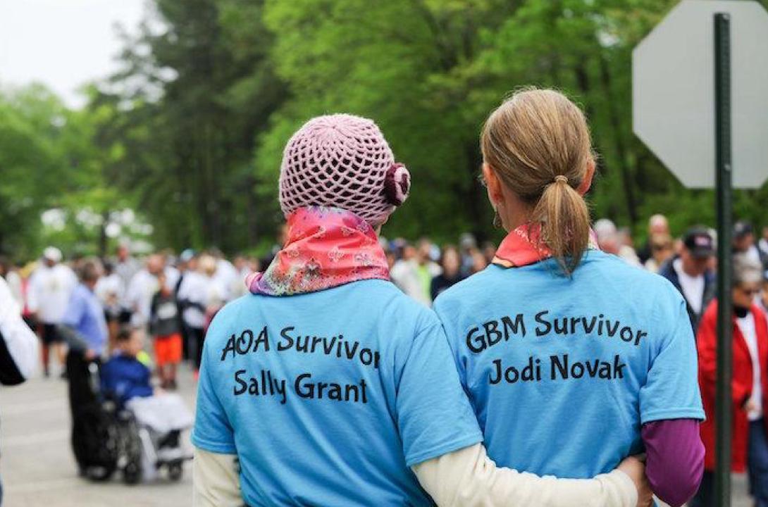 back view of two women in blue t-shirts facing a crowd of walkers. One is AOA Survivor Sally Grant and one is GBM survivor Jodi Novak