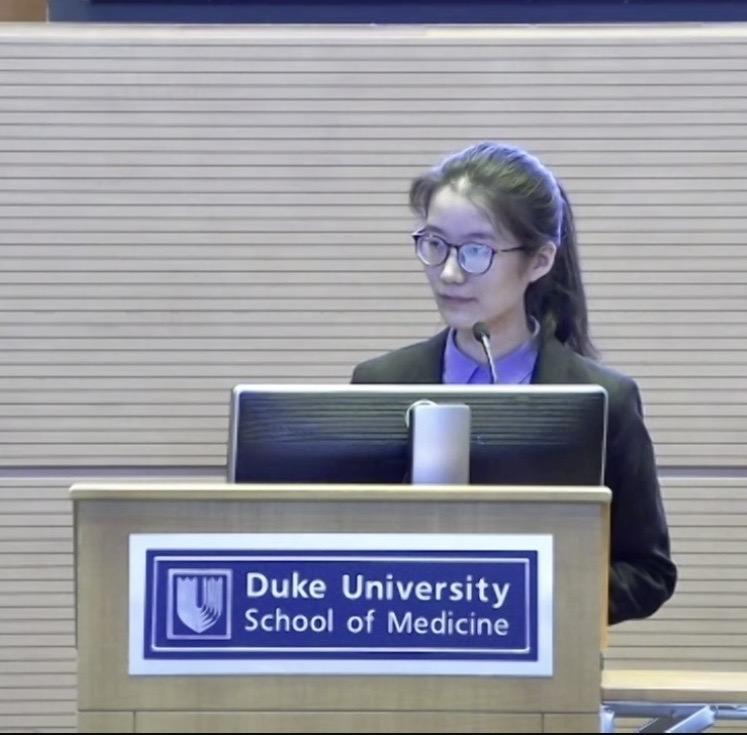 woman with glasses at podium labeled "Duke University School of Medicine"