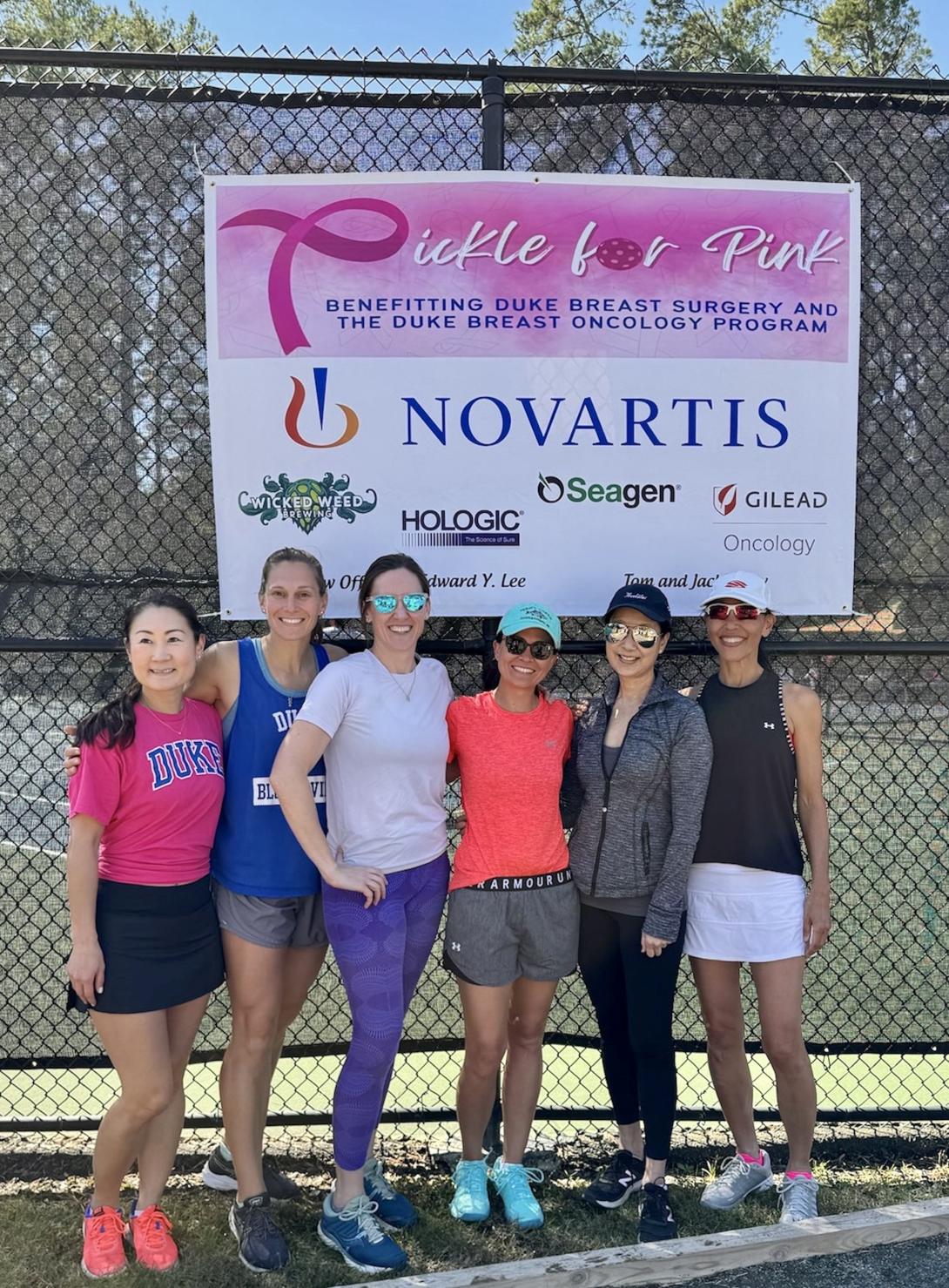 Six women in athleticwear pose in front of a black grated court under a "Pickle for Pink" event poster that includes a list of sponsors