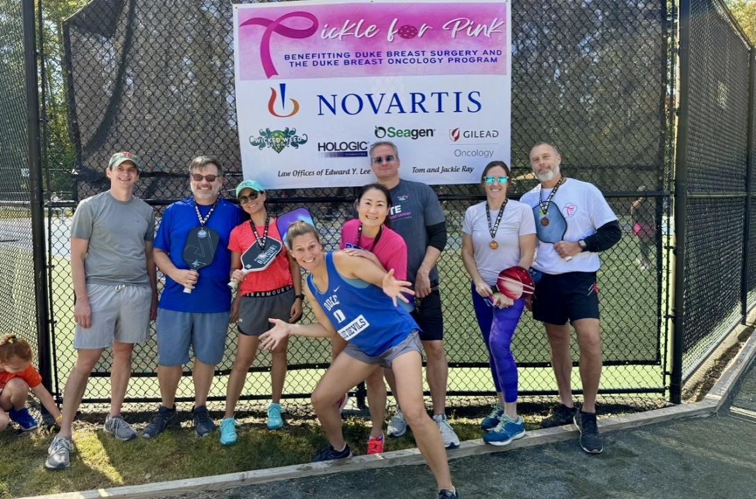 Eight men and women in athleticwear, most holding pickleball paddles, pose outside in front of a black grated court. A Pickle for Pink sign hangs above with a list of event sponsors.