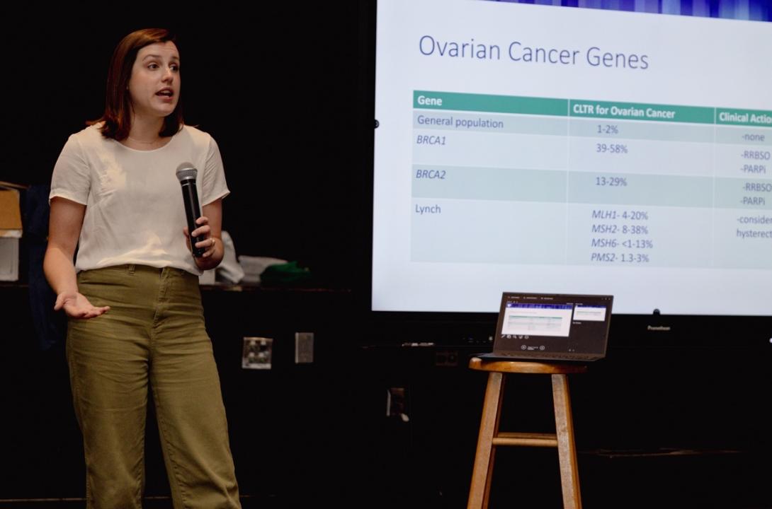 woman with a microphone standing near a poster that lists the various genetic drivers of ovarian cancer