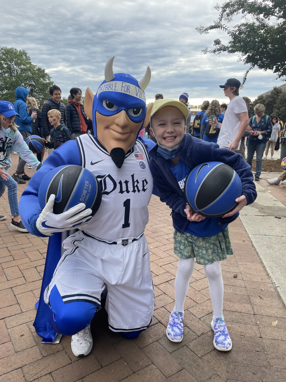 Blue Devil Mascot holding a basketball kneels next to small girl holding a basketball outdoors with several people in the background