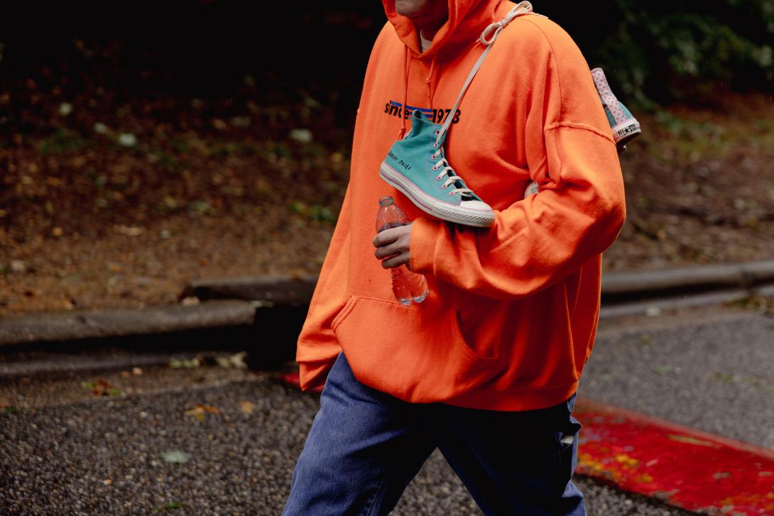 a man in an orange sweatshirt with teal sneakers tied together over his shoulder. On one sneaker is written "cancer sucks"
