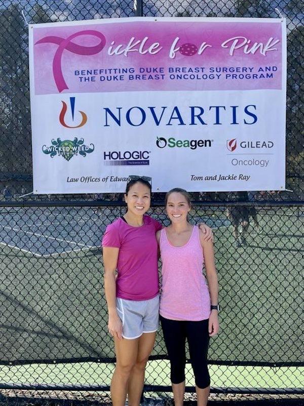 Two women dressed in pink stand under a "Pickle for Pink Sign" where the sponsors names and logos are written.