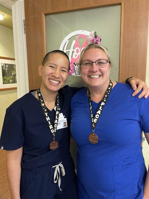 two women in scrubs pose with medals around their necks in front of a wood and glass door with a pink ribbon sticker that says "Hope, Never give up."