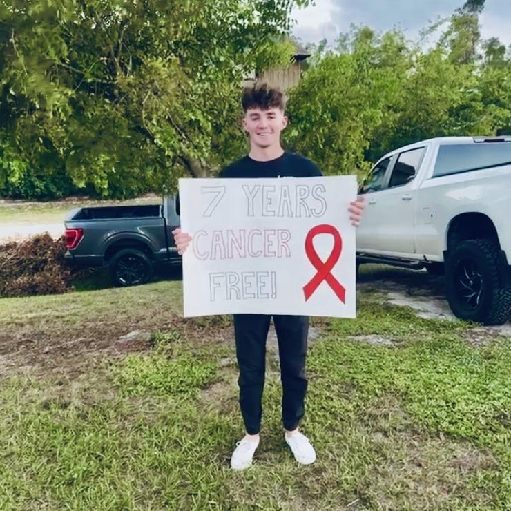 young man holding a sign in a yard that reads "7 Years Cancer Free" with a red ribbon