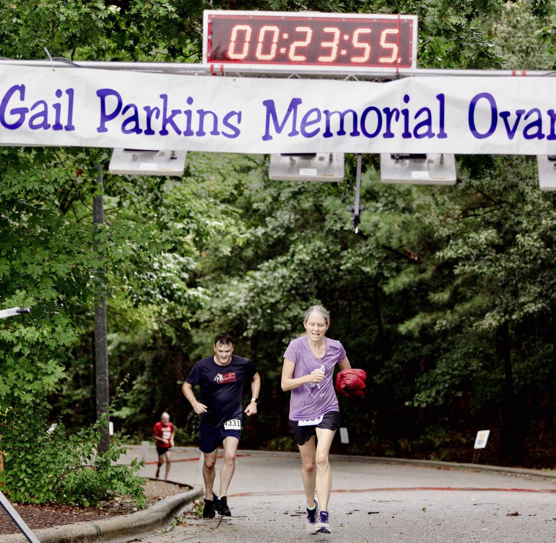 two runners, a man and a woman run toward the finish line beneath a Gail Parkins Memorial Ovarian banner and the time reading 00:23:55 