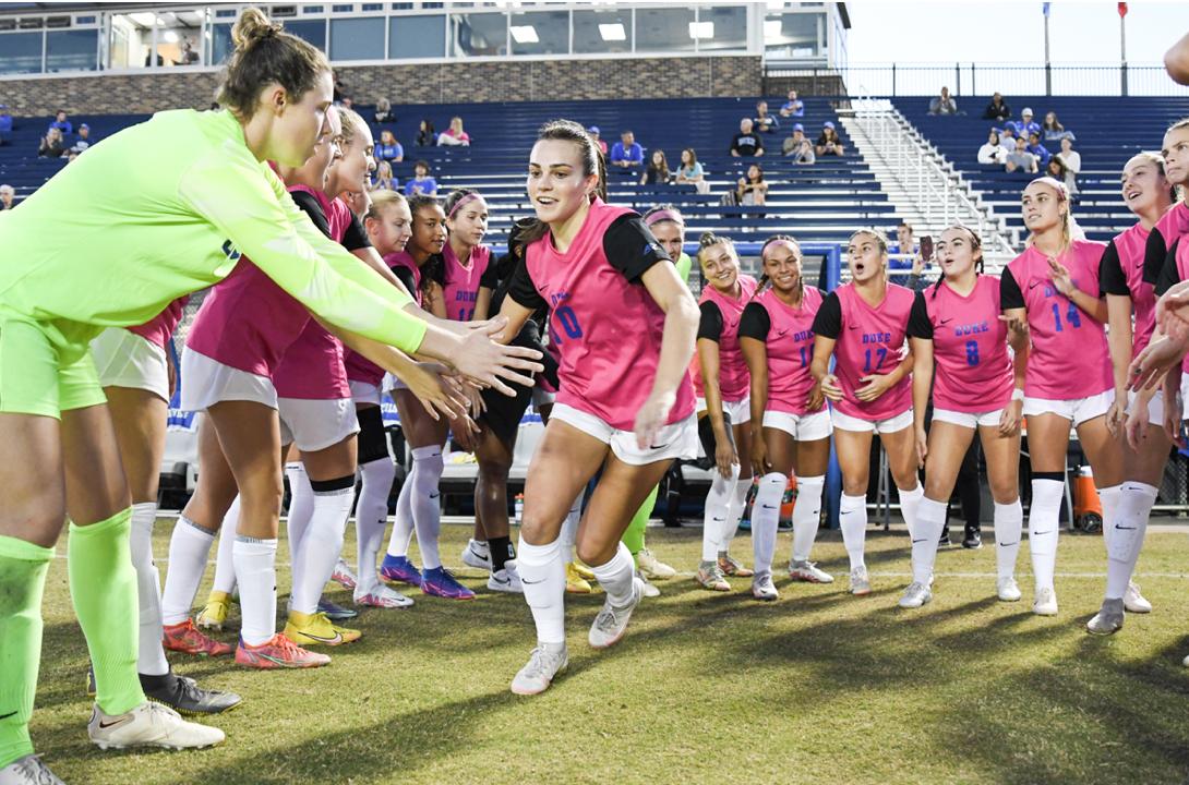 On the soccer field, 12 women in pink jerseys and white shorts surround one woman in the same uniform as she moves toward the goalie in green to shake hands